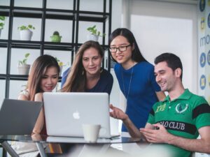 Four smiling people huddle around a laptop to review sales methods and techniques for their business.